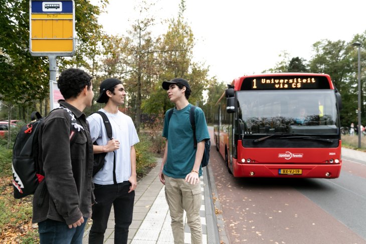 A group of three students waits at the bus stop. A bus drives up to them.