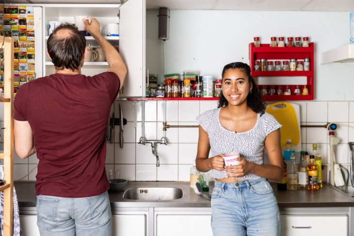 Two students in the kitchen of their student house. One is drinking a cup of coffee, the other is searching in a cabinet.